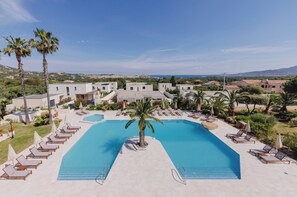 A resort with a large swimming pool, lounge chairs, palm trees, and a view of the surrounding landscape.