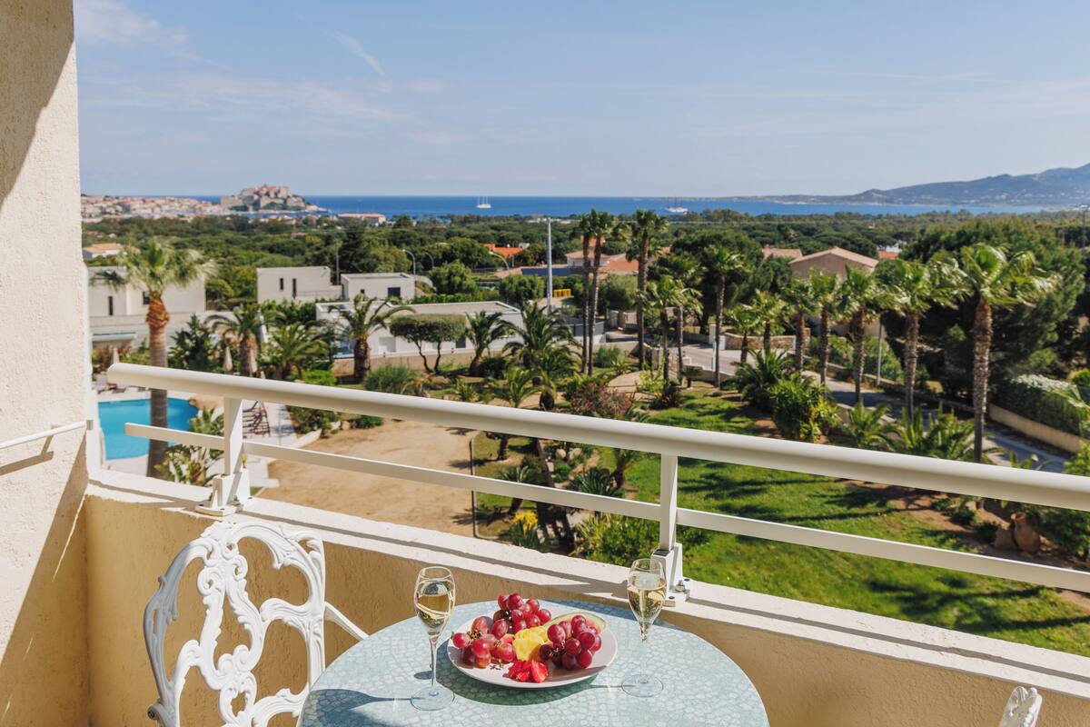 A balcony with a table, two glasses of white wine, and a plate of red grapes and a yellow fruit.