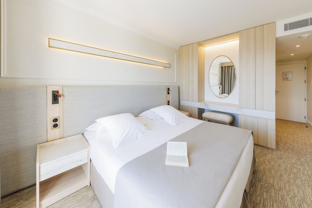 A hotel room with a bed, bedside tables, a desk, a mirror, and a wall-mounted light fixture.