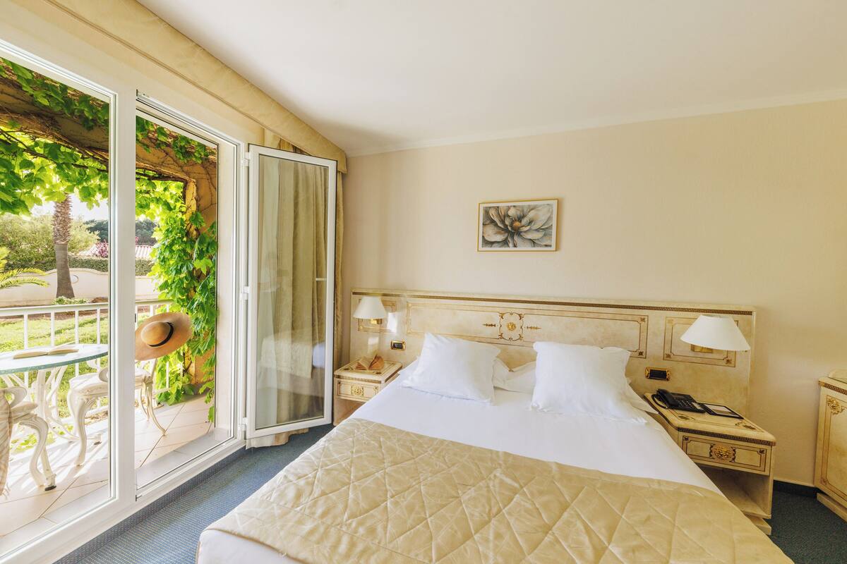 A hotel room with a bed, bedside tables, a nightstand, a window with a view of greenery, and a painting on the wall.