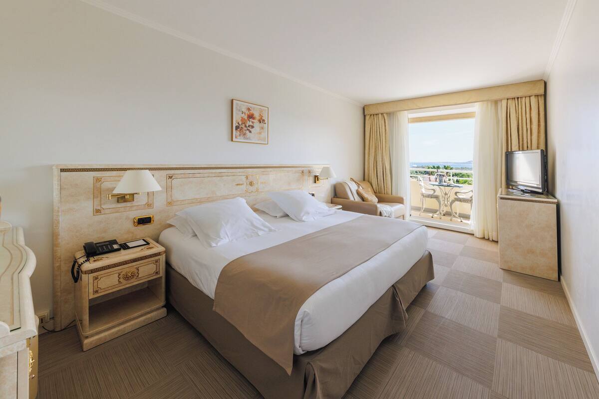 A hotel room with a large bed, a bedside table with a lamp, a TV, and a view of the outdoors.