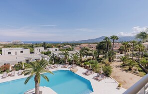 A resort with a swimming pool, lounge chairs, palm trees, and a view of the sea and mountains.