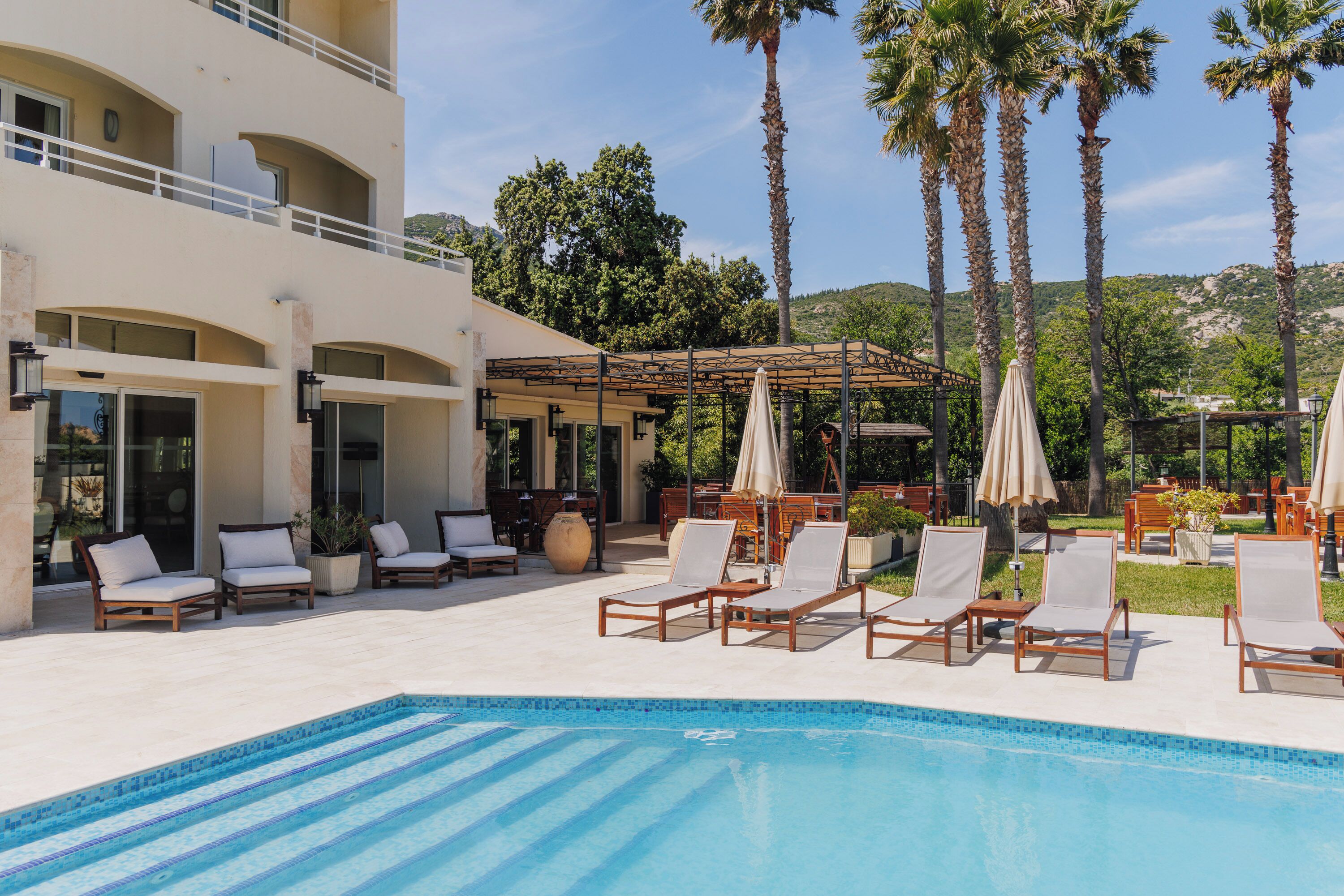 A pool area with lounge chairs and umbrellas, surrounded by palm trees and a building with multiple balconies.
