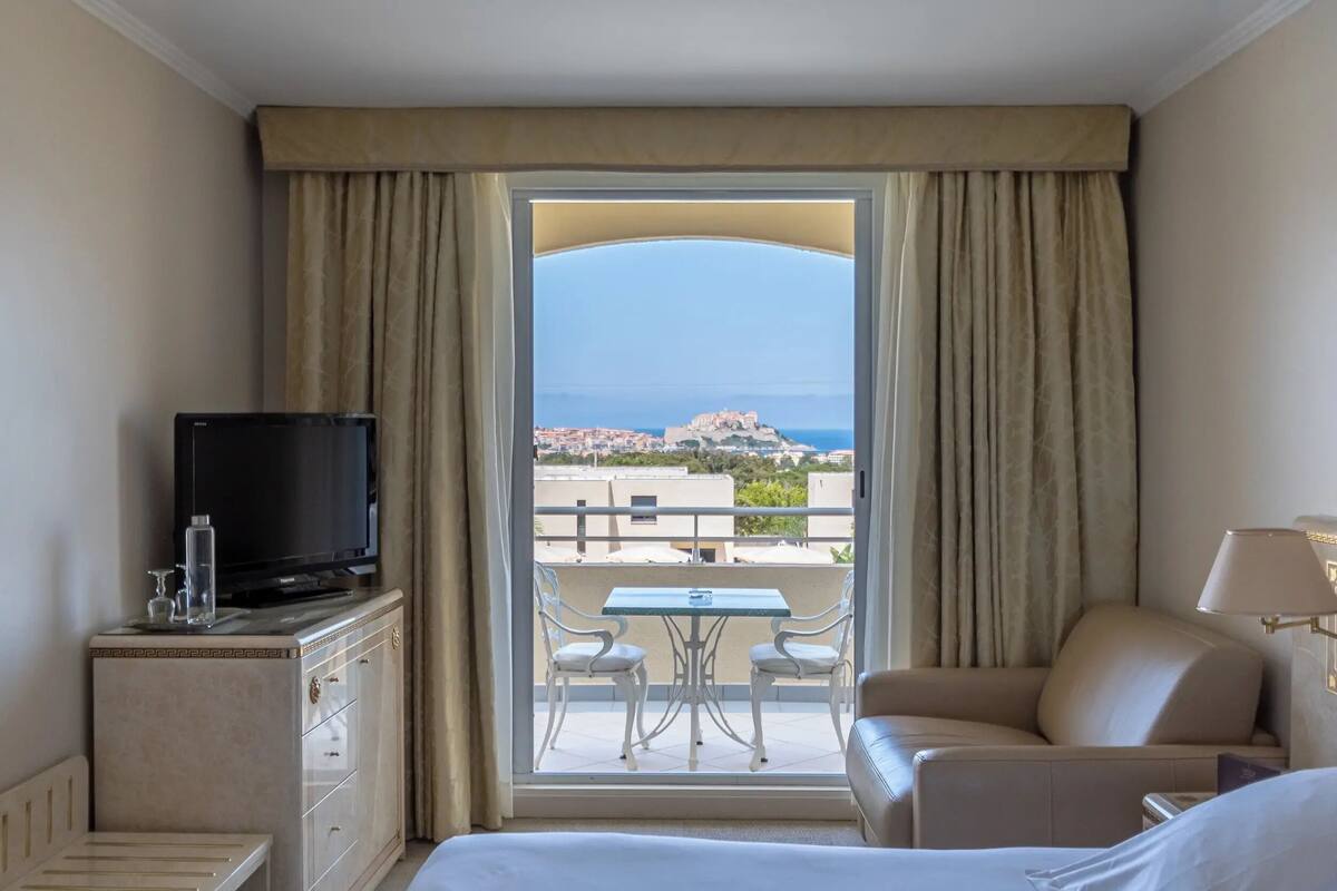 A hotel room with a balcony, a television, a table with chairs, and a sofa.