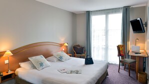 Ein Hotelzimmer mit Bett, Nachttischlampen, einem Schreibtisch mit Laptop, einem Sessel und einem Fernseher.