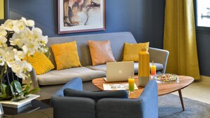 Ein modernes Wohnzimmer mit einem grauen Sofa, gelben Kissen, einem Couchtisch aus Holz mit einem Laptop, einem Glas Orangensaft und einer Schüssel bunte Cerealien.