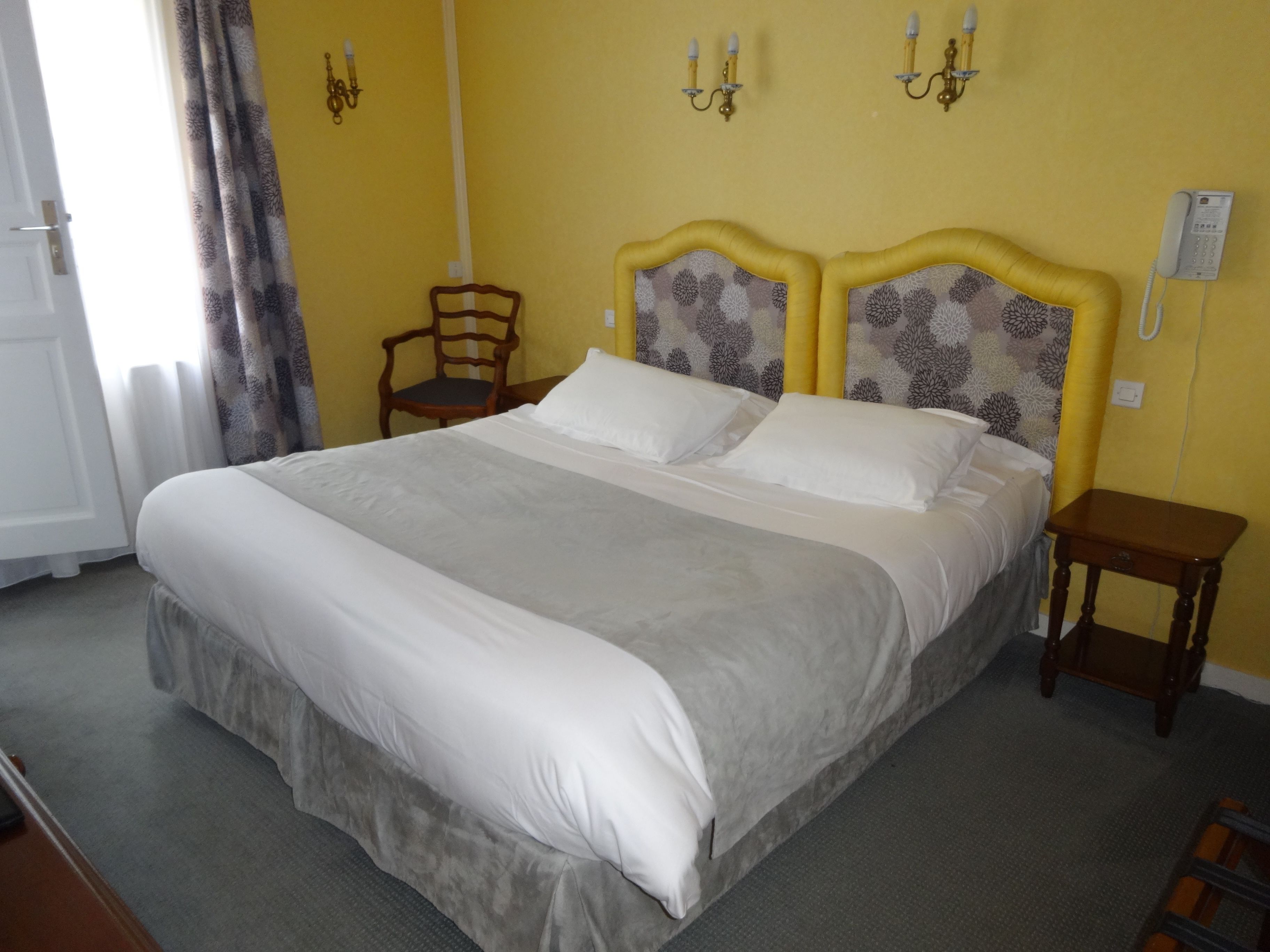 A neatly made bed with a grey and white duvet, a wooden chair, and a bedside table with a phone.