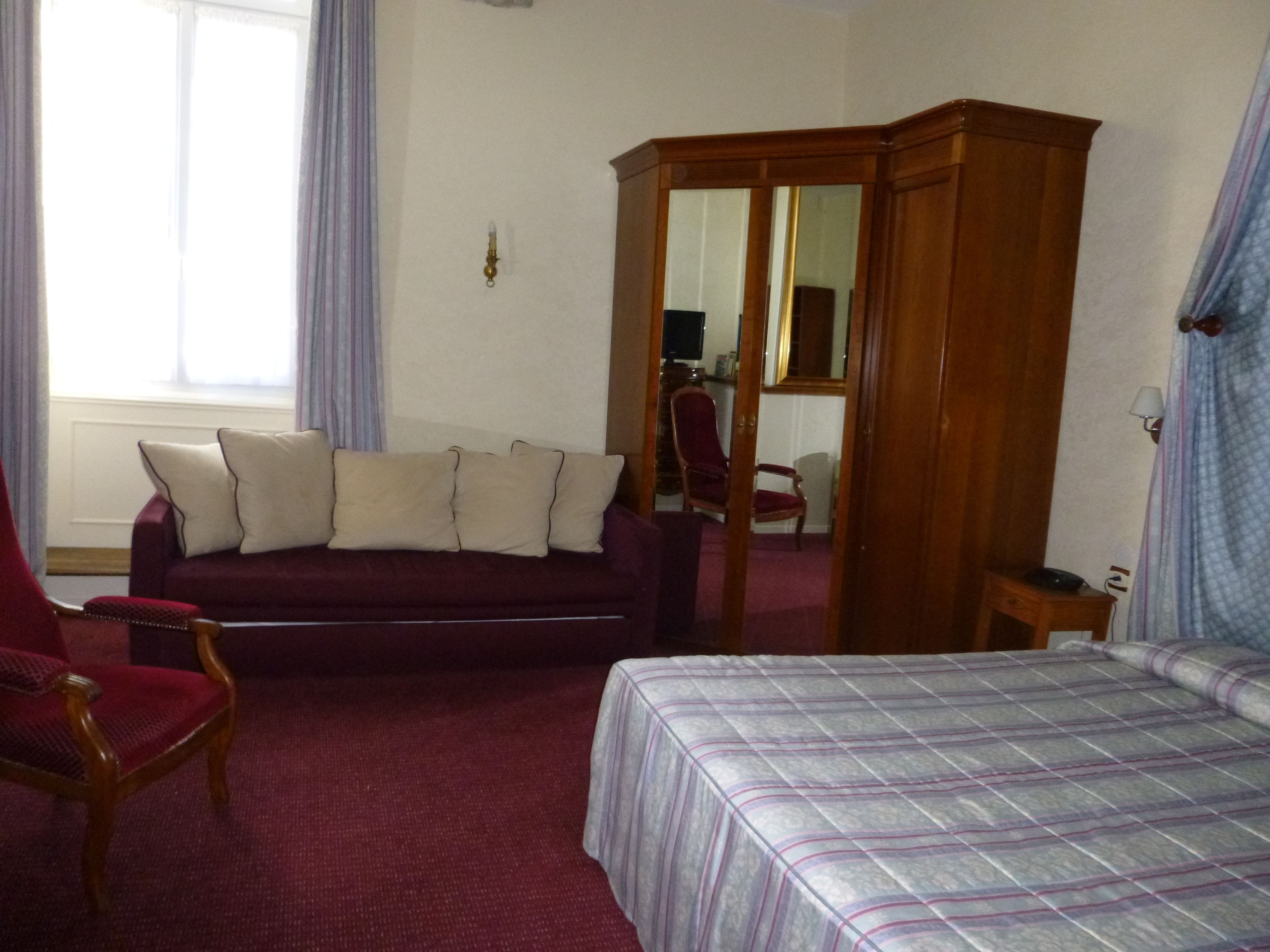 A room with a red carpet, a red armchair, a dark red sofa with pillows, a wooden wardrobe, and a bed with a striped bedspread.