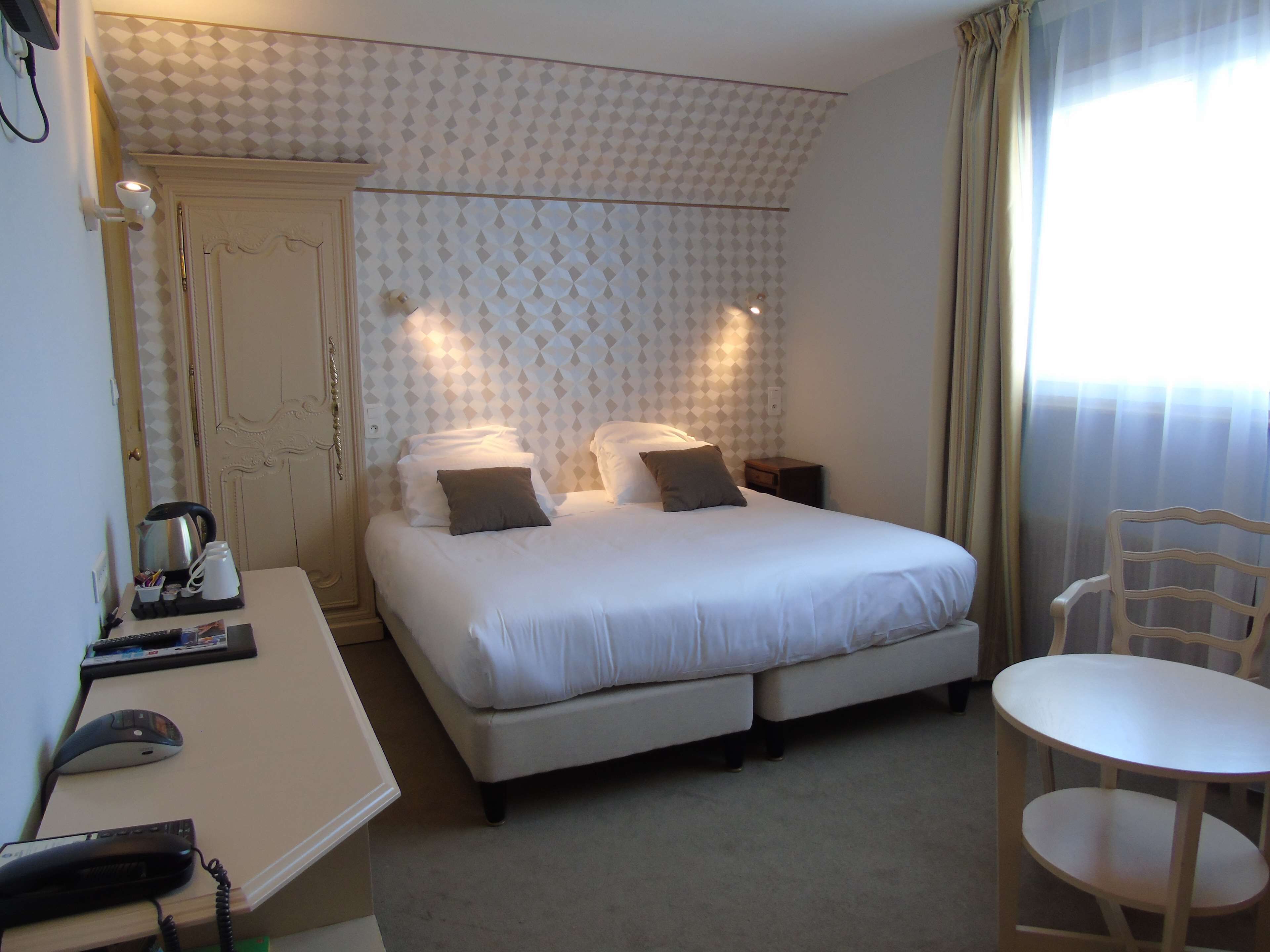 A hotel room with a bed, bedside table, chair, and a desk with a telephone and a kettle.