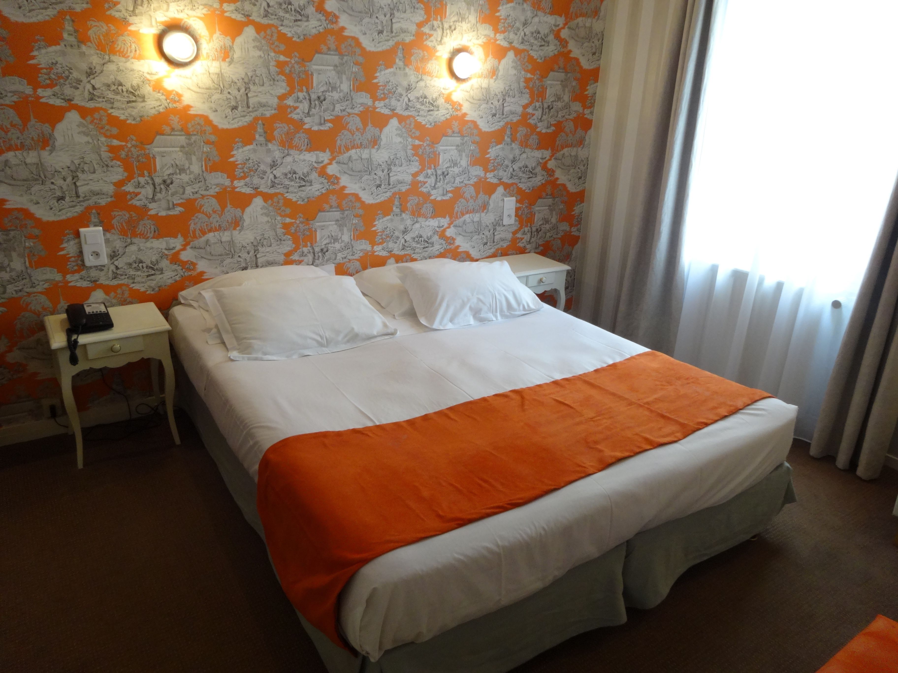 A hotel room with a bed, white pillows, an orange blanket, a nightstand with a phone, and a wall with a patterned wallpaper.