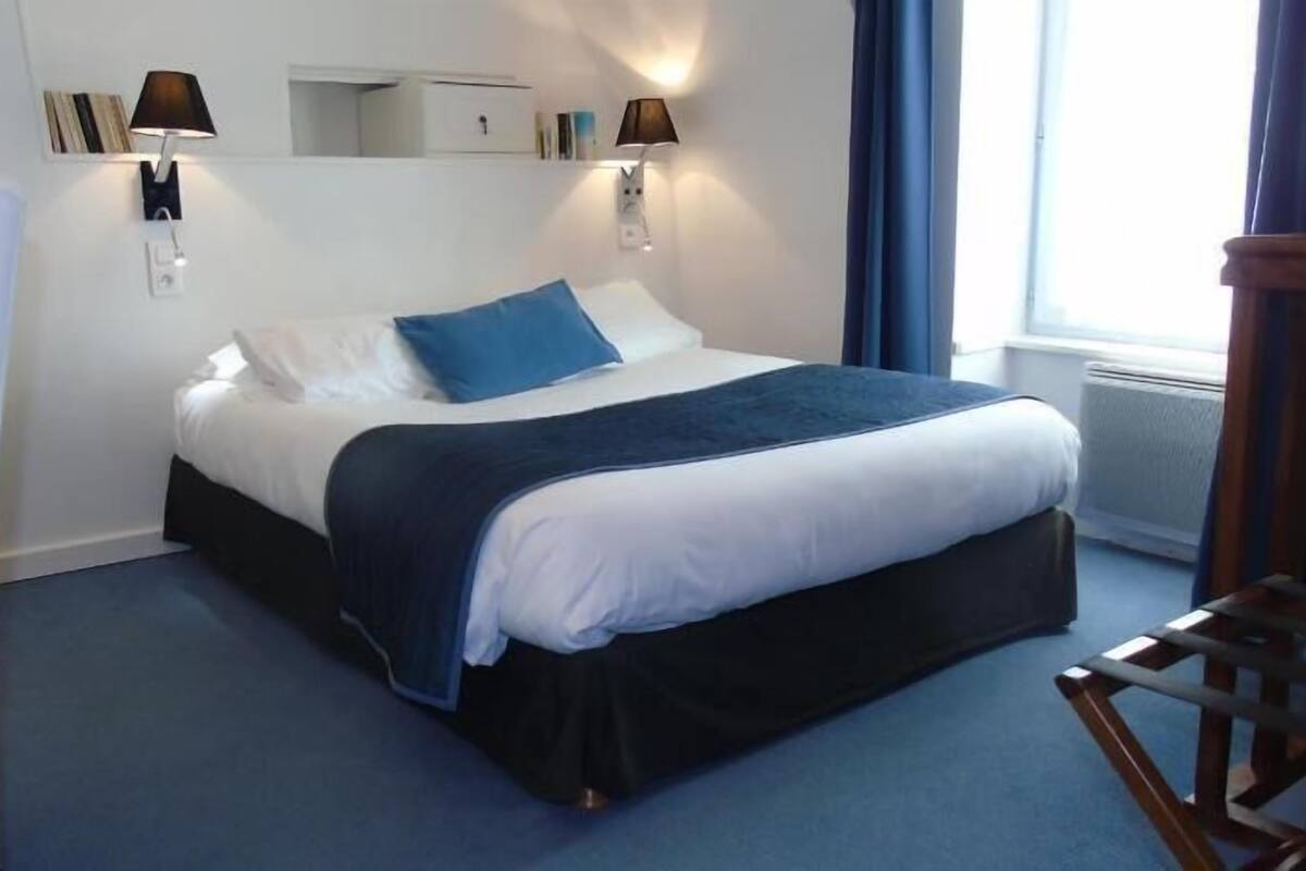 A bed with a blue blanket and pillows, a bedside table, a chair, a wall-mounted lamp, and a window with curtains.