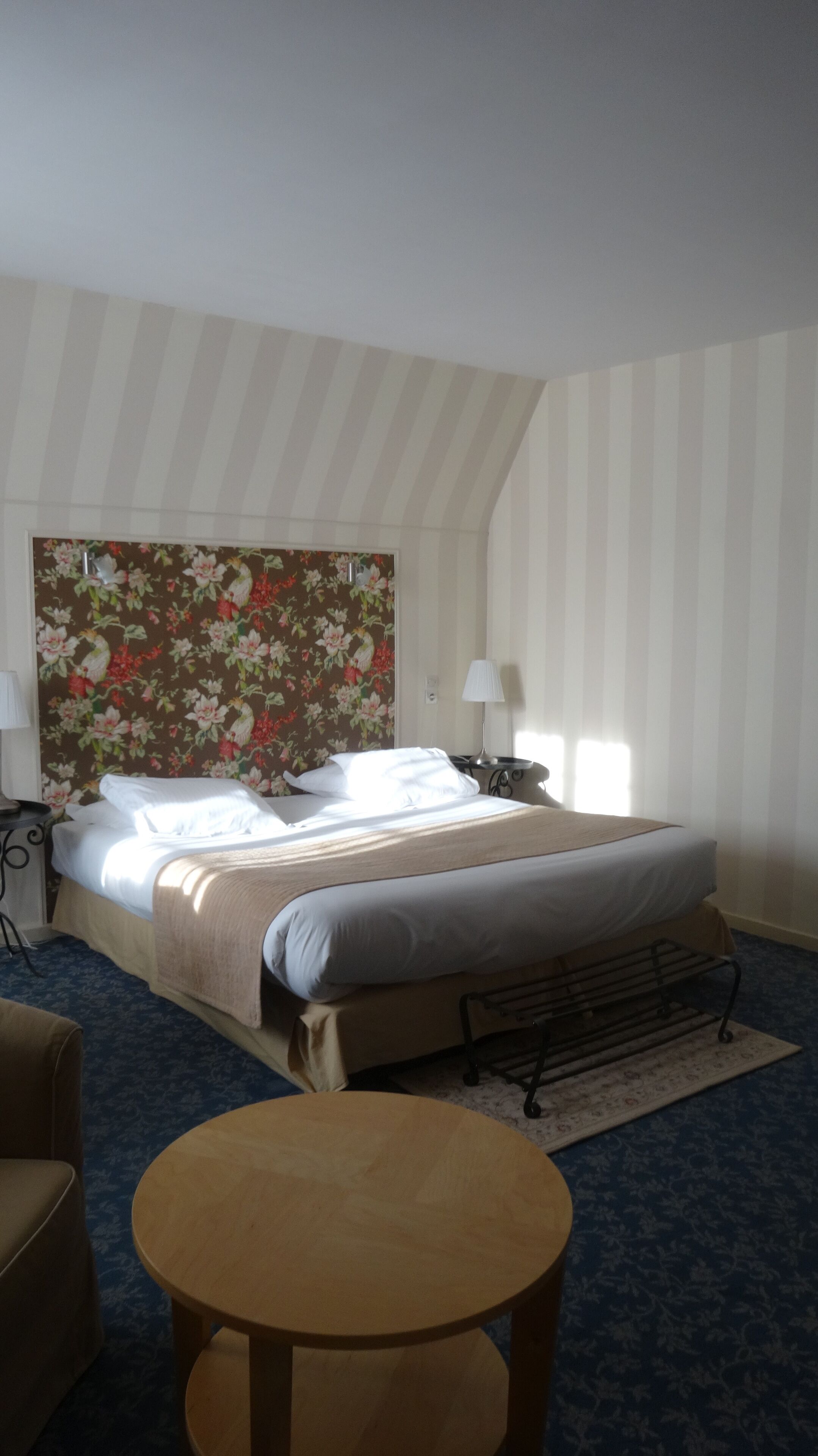 A hotel room with a bed, a bedside table, a lamp, a chair, and a small round table.