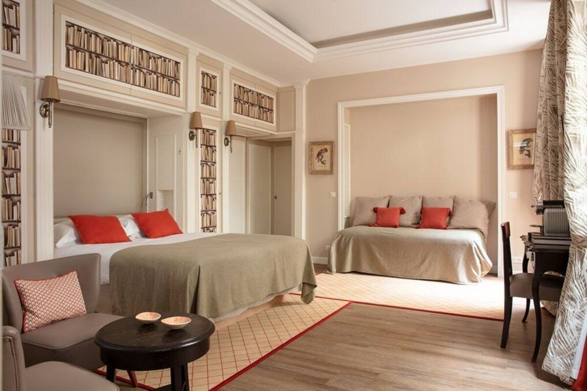 A hotel room with two beds, a sofa, a small table, and a bookshelf.