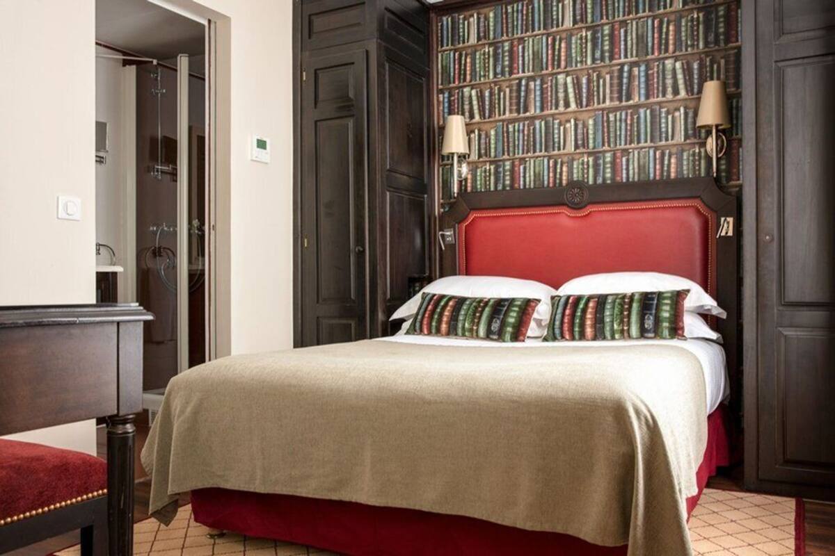 A hotel room with a large bed, a bookshelf, and a red upholstered chair.