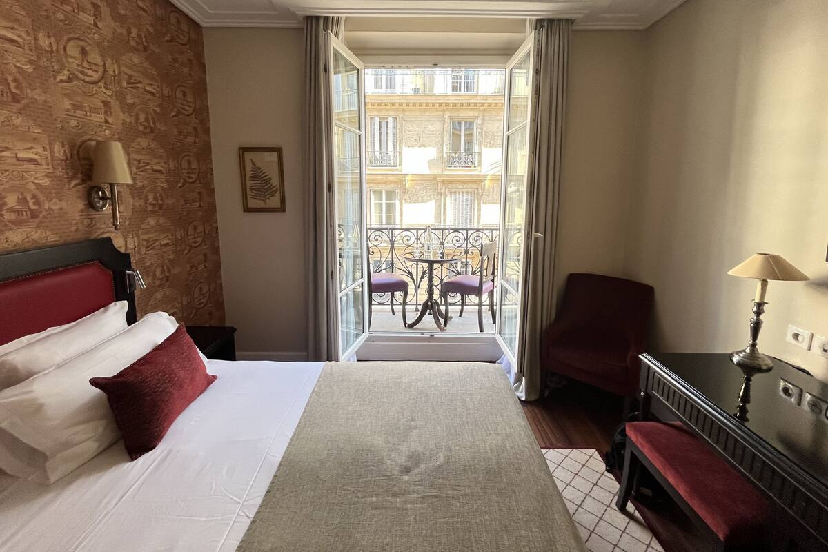 A hotel room with a bed, a desk, a chair, a balcony with a table and chairs, and a view of the building outside.
