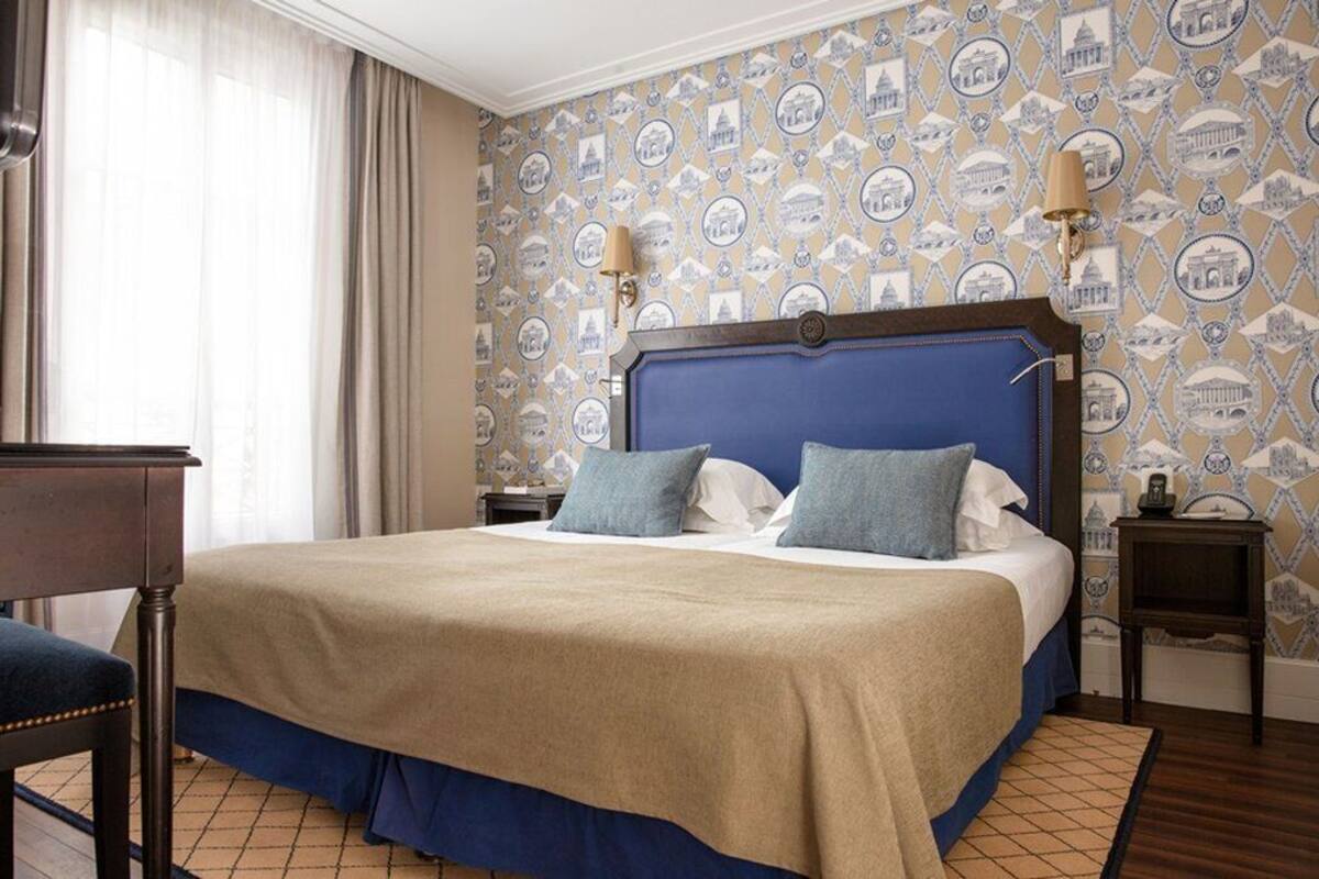 A hotel room with a large bed, a wooden nightstand, a desk, and a patterned wallpaper.