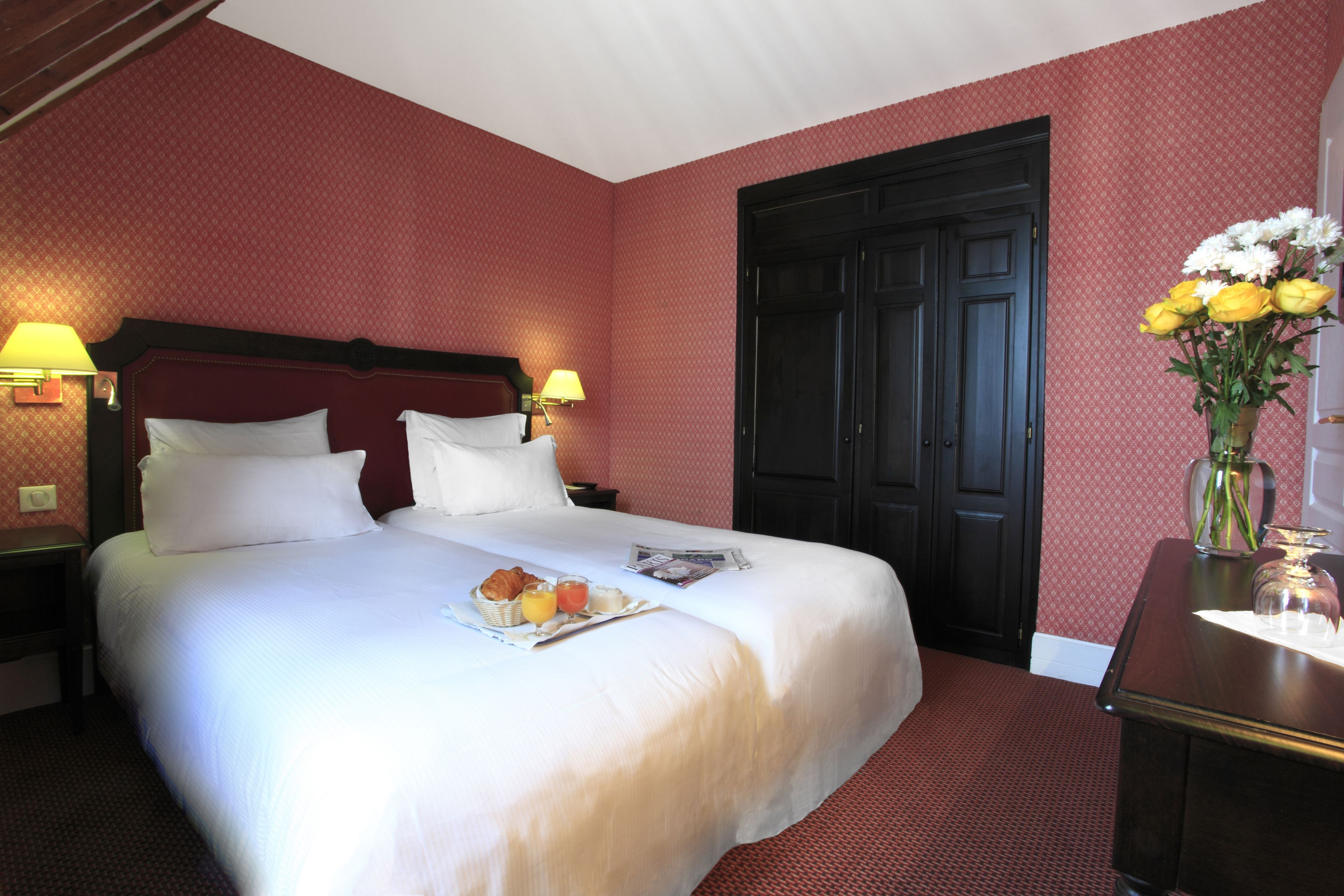 Une chambre d’hôtel avec un grand lit, deux tables de chevet avec des lampes, une porte en bois sombre et un vase rempli de fleurs posé sur une commode.