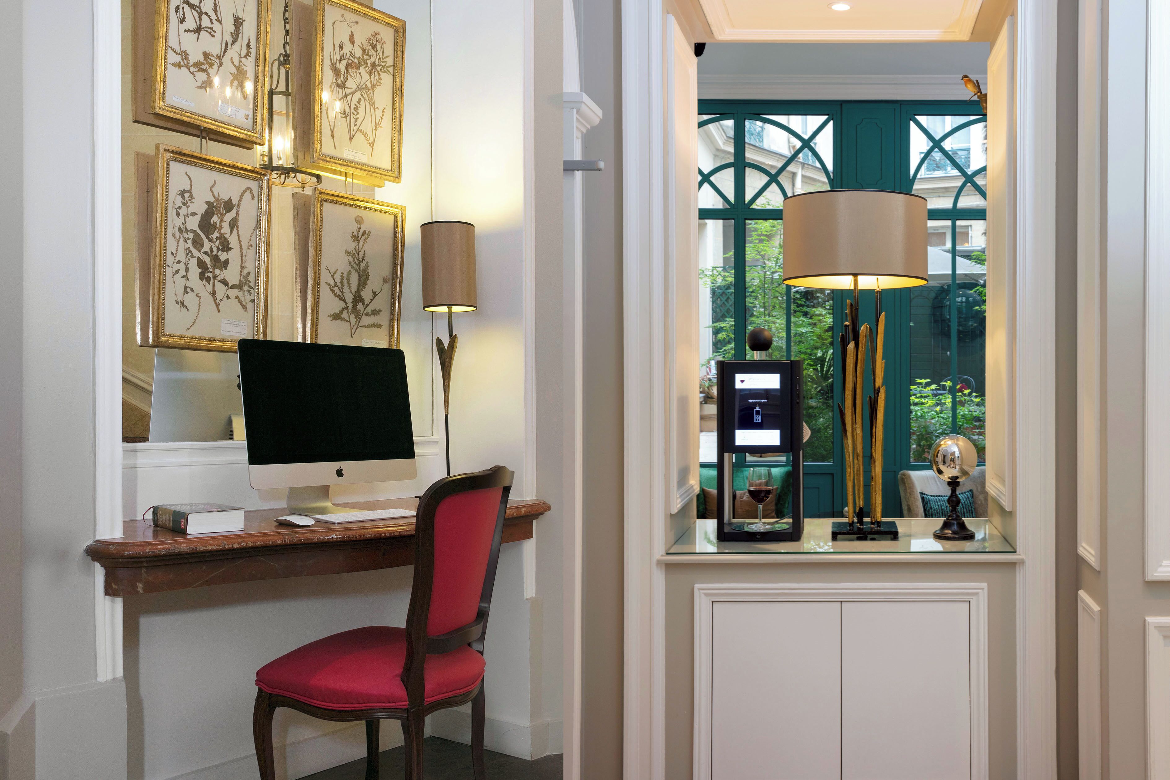A study with a desk, chair, and a computer monitor. There are framed botanical prints on the wall and a window with a view of greenery.