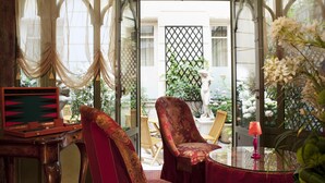 A room with a view of a garden, a mahjong table, two ornate chairs, and a small table with a pink lamp.