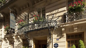 A hotel entrance with a wrought iron balcony, a sign indicating the hotel name, and a blue sign with a white 'H' symbol.