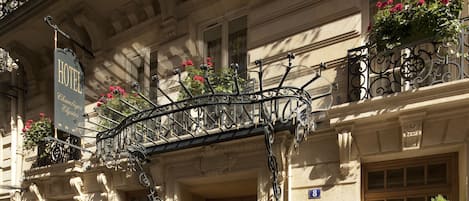 A hotel entrance with a wrought iron balcony, a sign indicating the hotel name, and a blue sign with a white 'H' symbol.