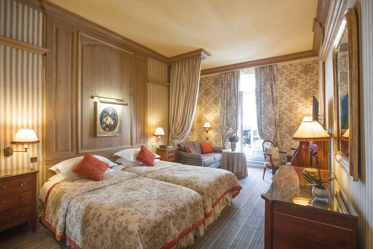 A hotel room with two beds, a sitting area with a sofa and armchairs, a desk with a lamp, and a large mirror.