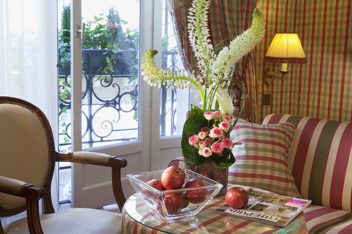 A room with a glass table holding a bowl of apples, a vase with flowers, and a magazine.