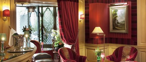 A luxurious room with a red and gold color scheme, featuring a large window with curtains, a painting on the wall, and a seating area with a table and chairs.