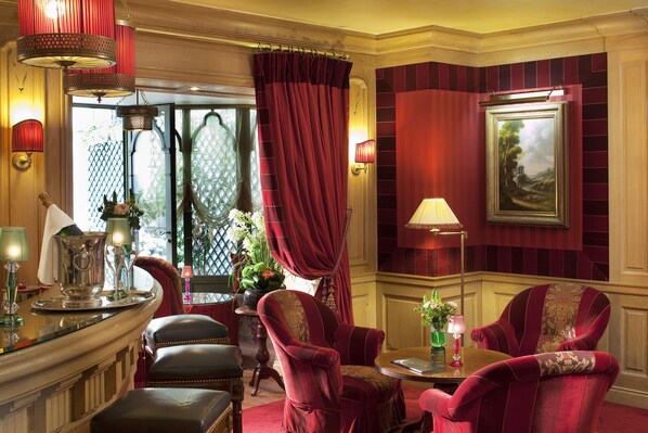 A luxurious room with a red and gold color scheme, featuring a large window with curtains, a painting on the wall, and a seating area with a table and chairs.