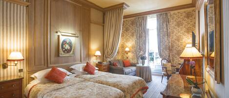 Habitación de hotel con dos camas, zona de estar con sofá y sillones, escritorio con lámpara y un espejo grande.