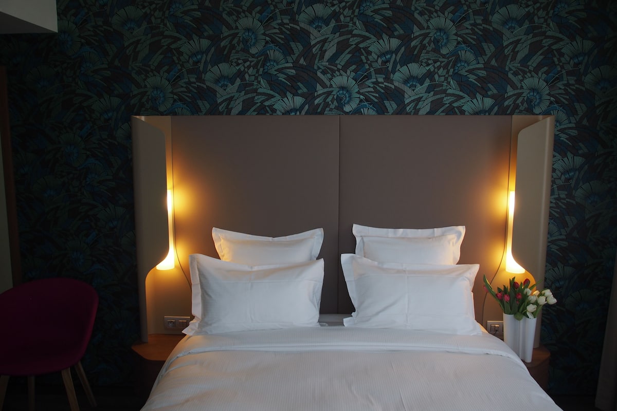 Habitación de hotel con una cama grande, dos lámparas de noche, una silla roja y un papel tapiz con motivos florales.