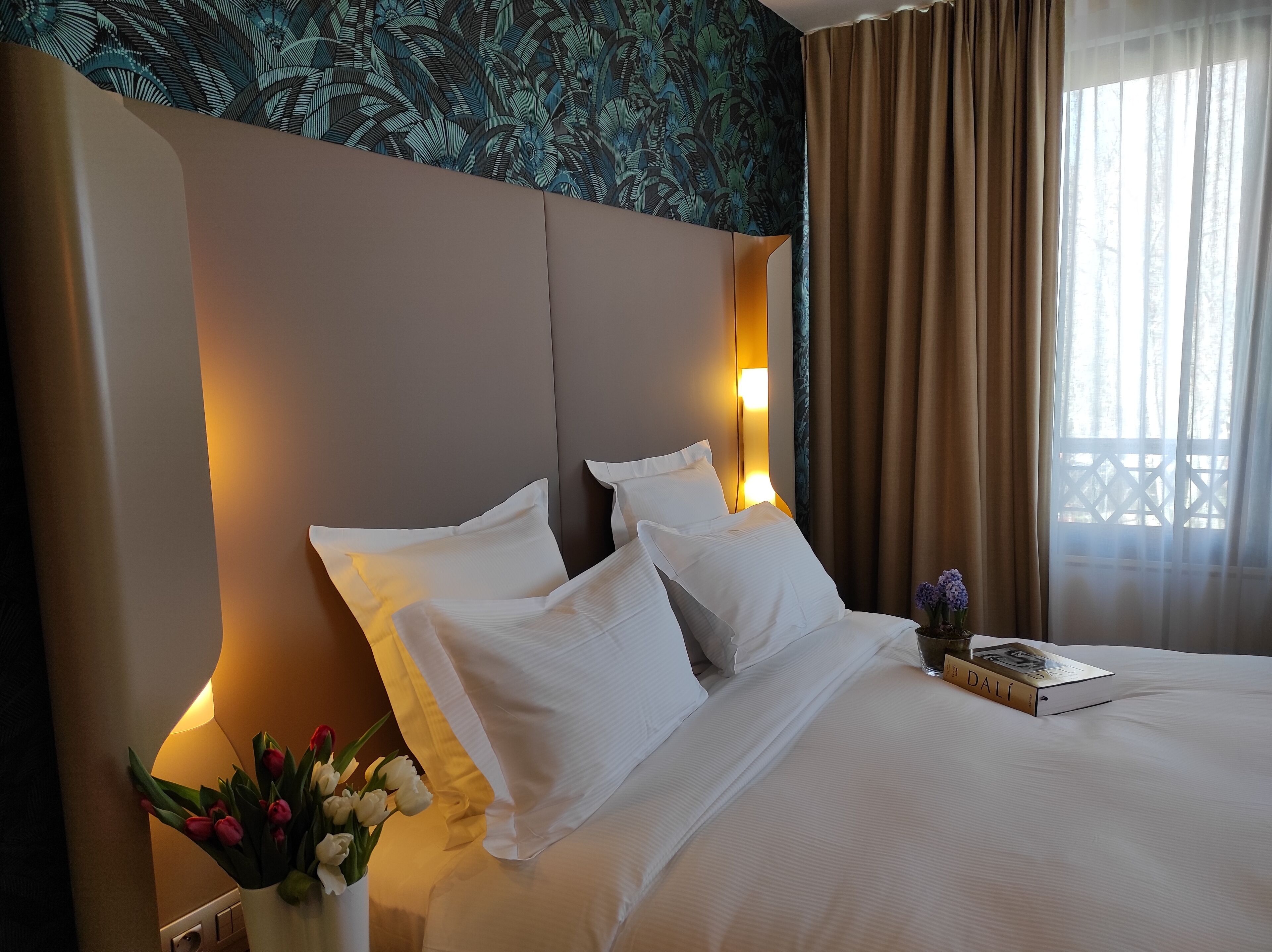 Habitación de hotel con una cama grande, ropa de cama blanca, un florero con flores, un libro y una ventana con cortinas.
