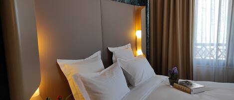 Habitación de hotel con una cama grande, ropa de cama blanca, un florero con flores, un libro y una ventana con cortinas.