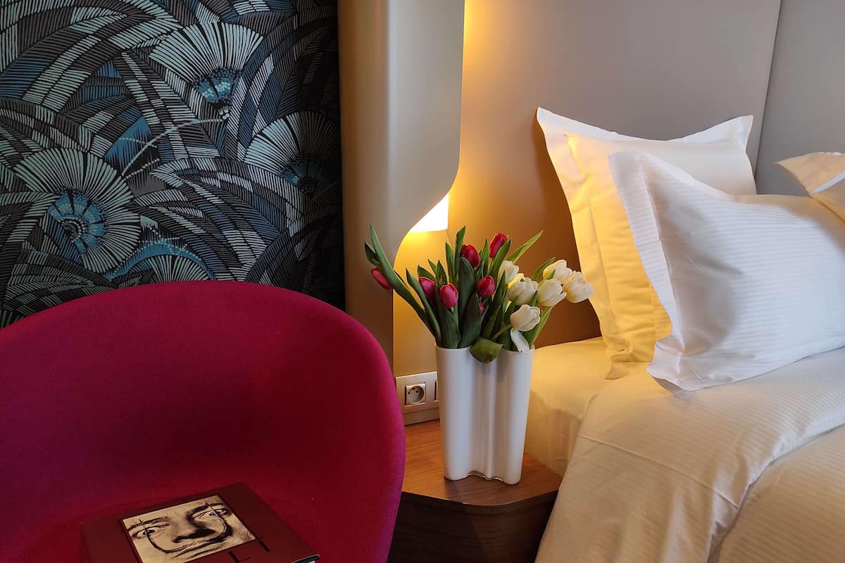 Una habitación de hotel con una silla roja, una mesita de noche con un libro, un florero con flores y una cama con almohadas blancas.