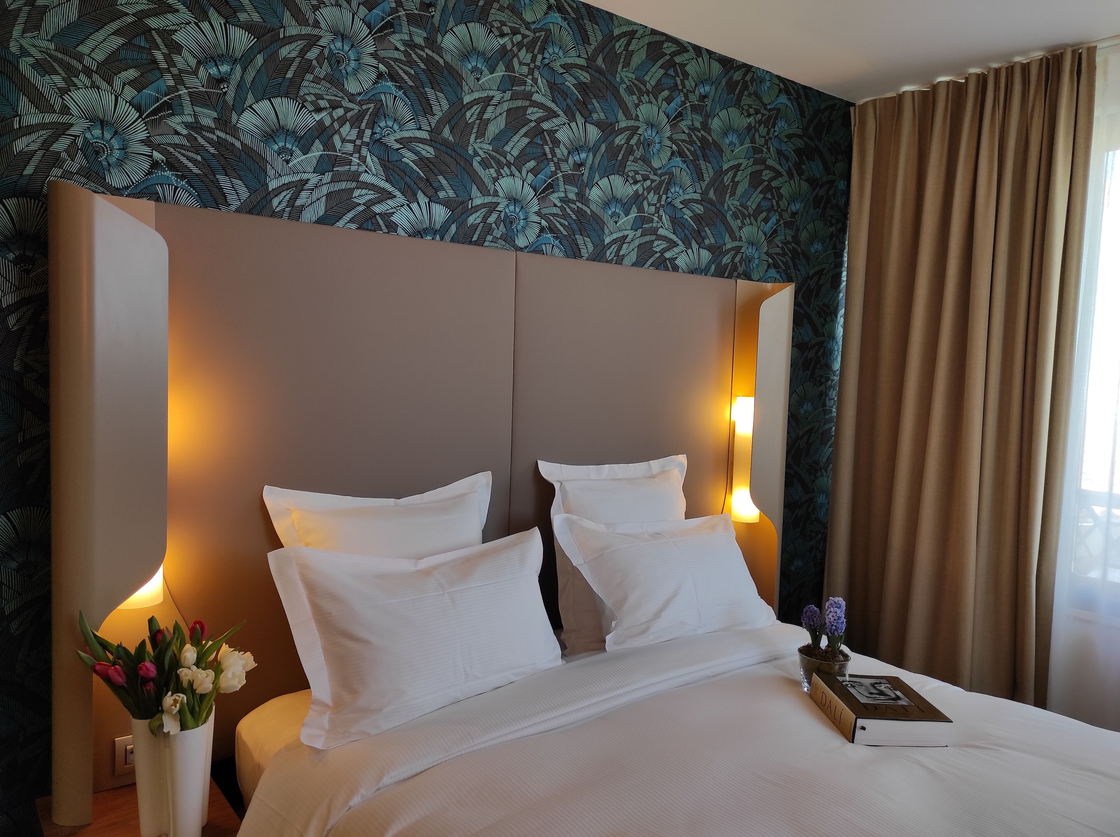 A hotel room with a large bed, white linens, a headboard with integrated lighting, a vase with flowers, and a book on the bed.