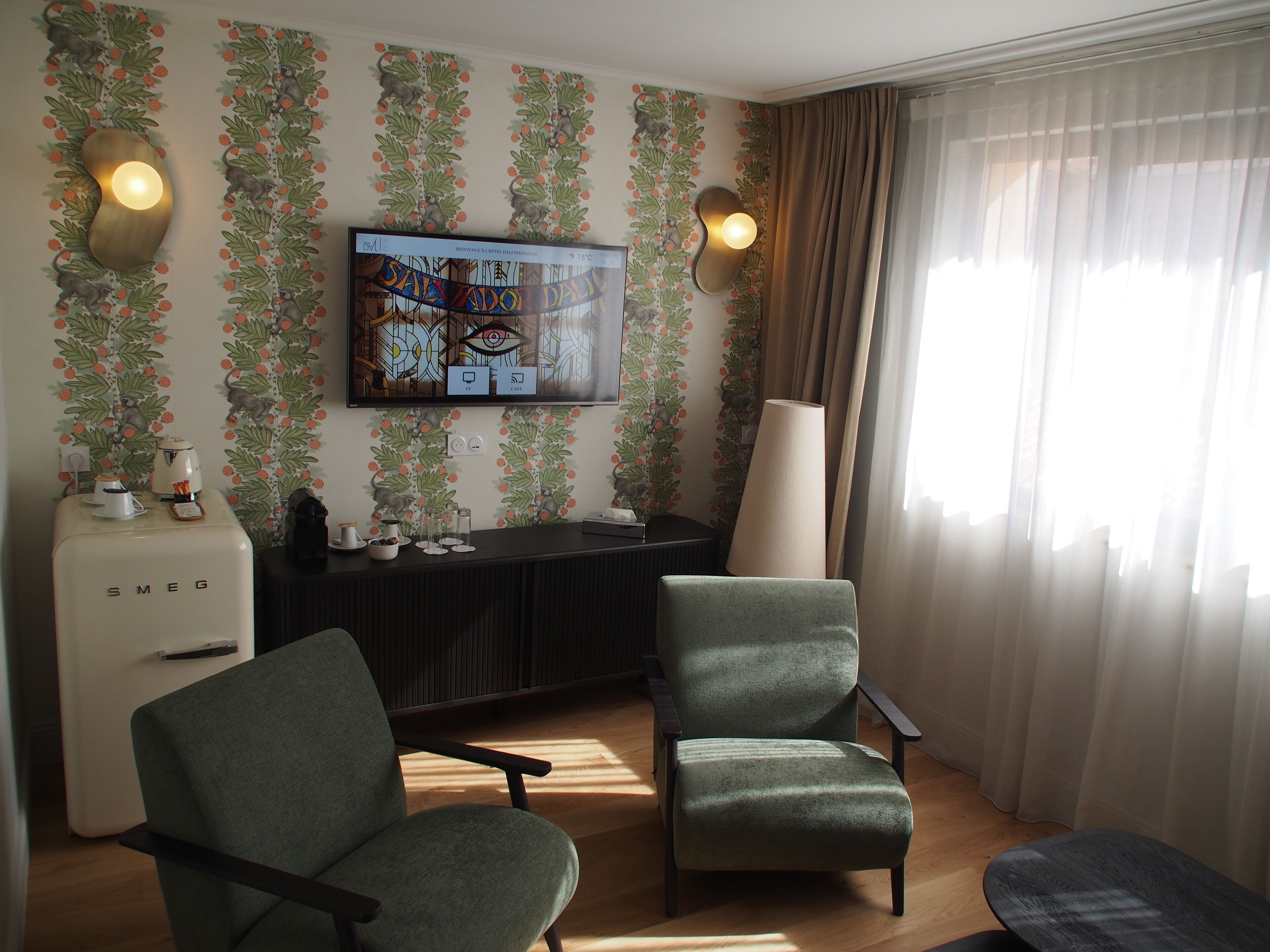 Una sala de estar con un televisor de pantalla plana montado en la pared, dos sillones, una cafetera y una lámpara.