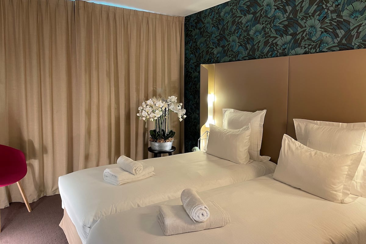 Habitación de hotel con una cama grande, dos almohadas, una mesita de noche con una lámpara, un jarrón con flores blancas y una silla roja.