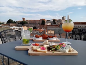 A table with a variety of food items and a drink, set against a backdrop of a cityscape with historical buildings. - Dali Hôtel - Restaurant (Perpignan)