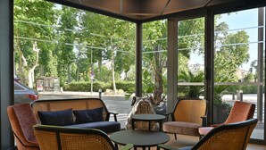 A modern indoor seating area with wicker furniture, large windows offering a view of the street and greenery outside.