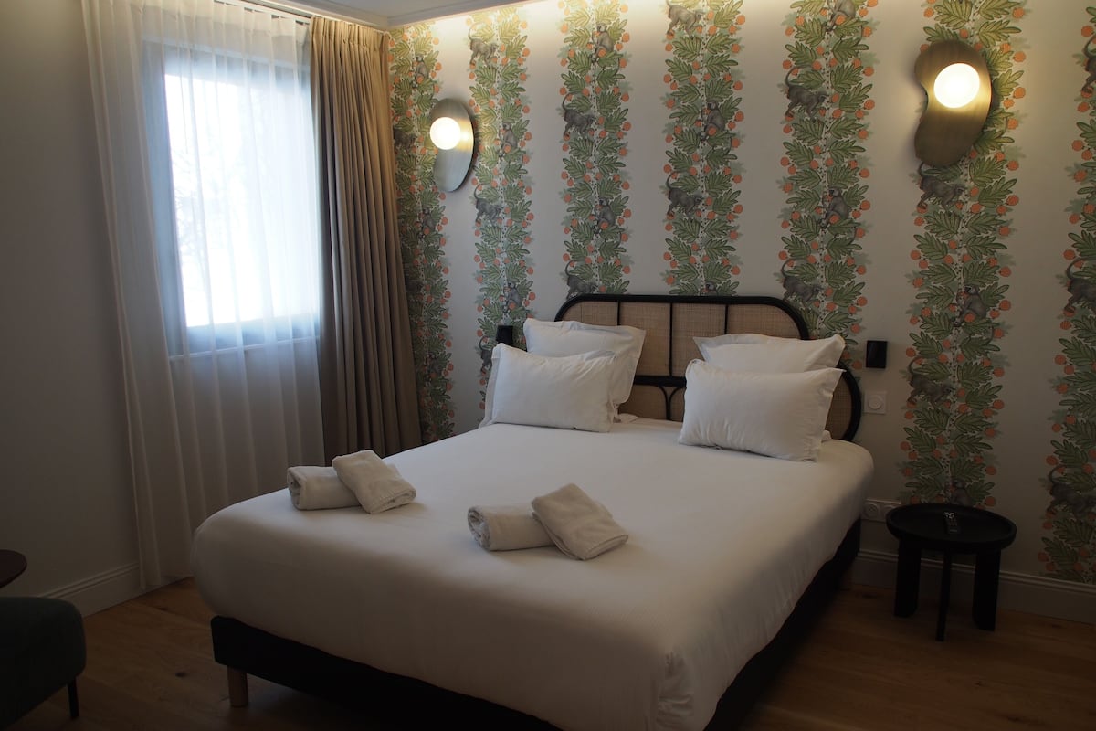 Una cama bien hecha con sábanas blancas y dos almohadas, una mesita de noche con una lámpara, y una pared con un estampado floral.