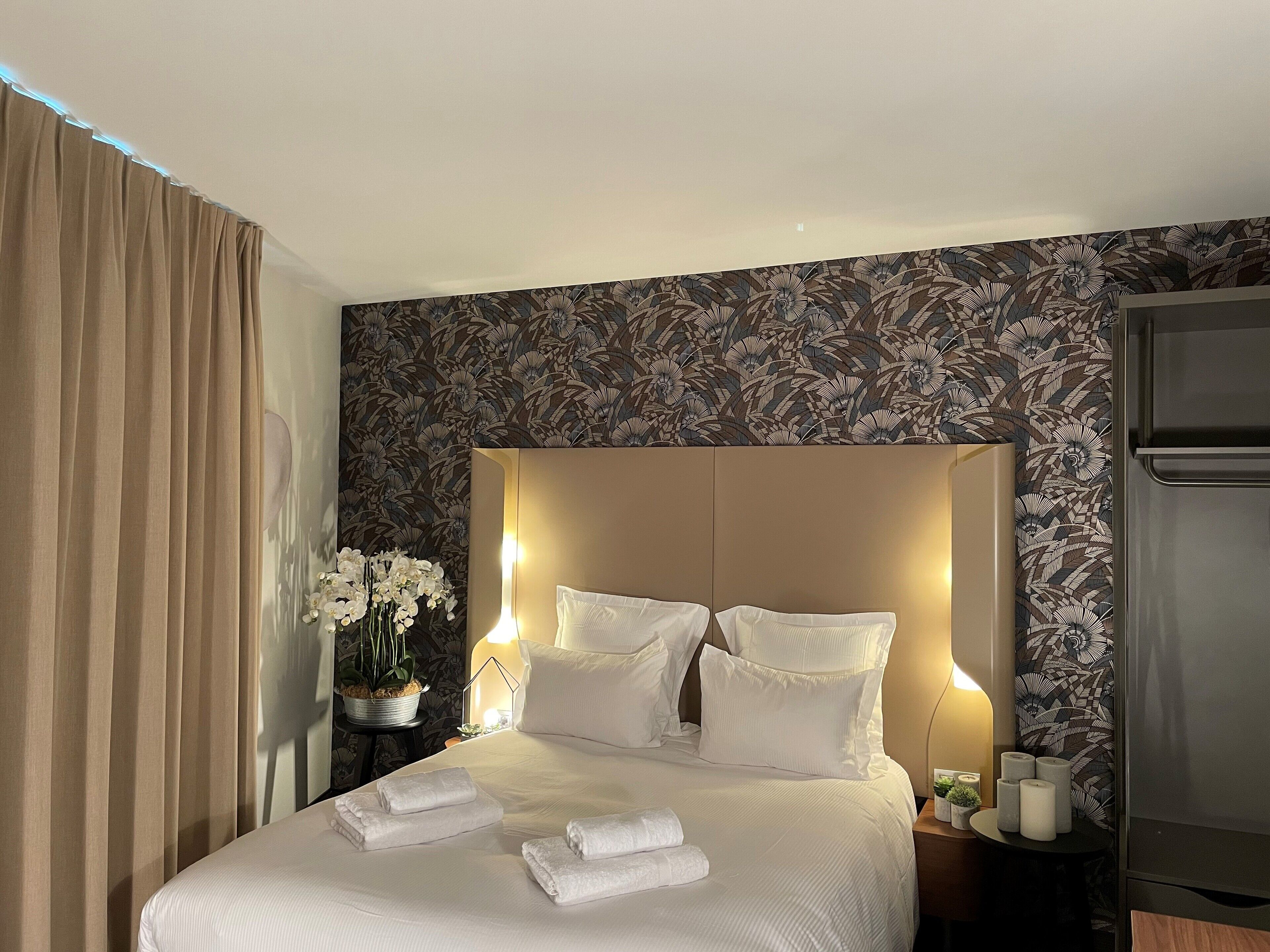 A hotel room with a large bed, white linens, a headboard with integrated lighting, a small table with a vase of flowers, and a refrigerator.