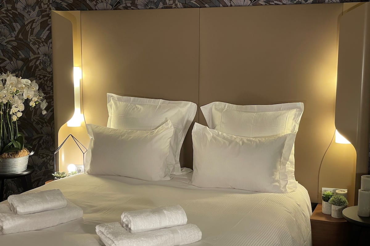 Una cama bien hecha con sábanas y almohadas blancas, una mesita de noche con una lámpara y una orquídea en maceta.