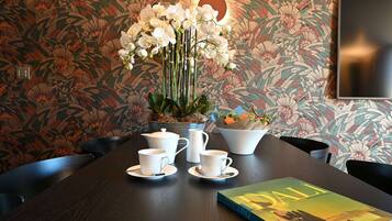 Een eettafel met witte kopjes, schoteltjes en een karaf, voor een behang met bloemen.
