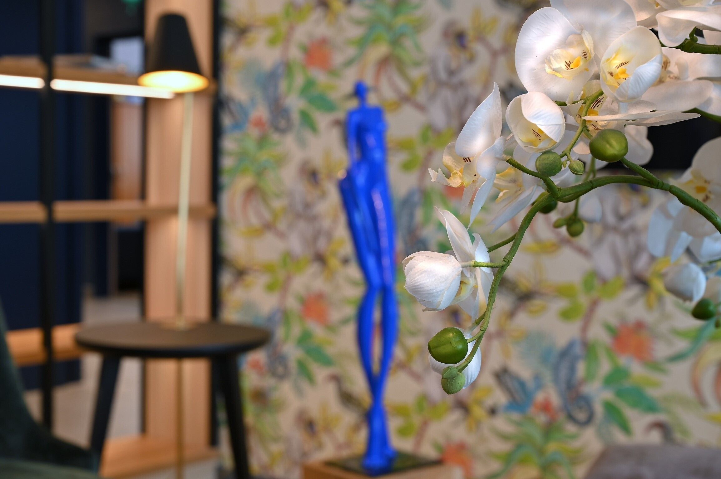 A close-up of white orchid flowers with green buds, a blue sculpture, and a patterned wallpaper background.