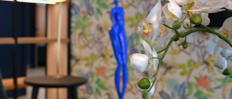 A close-up of white orchid flowers with green buds, a blue sculpture, and a patterned wallpaper background.