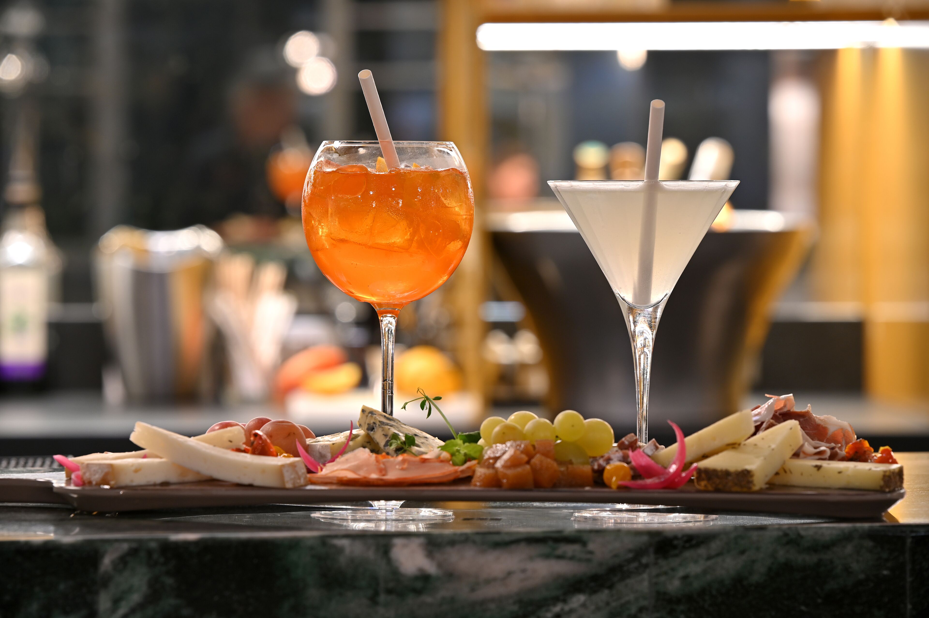 En bardisk med ett martiniglas och ett cocktailglas med sugrör, samt ett urval av ostar och charkuterier på en tallrik.