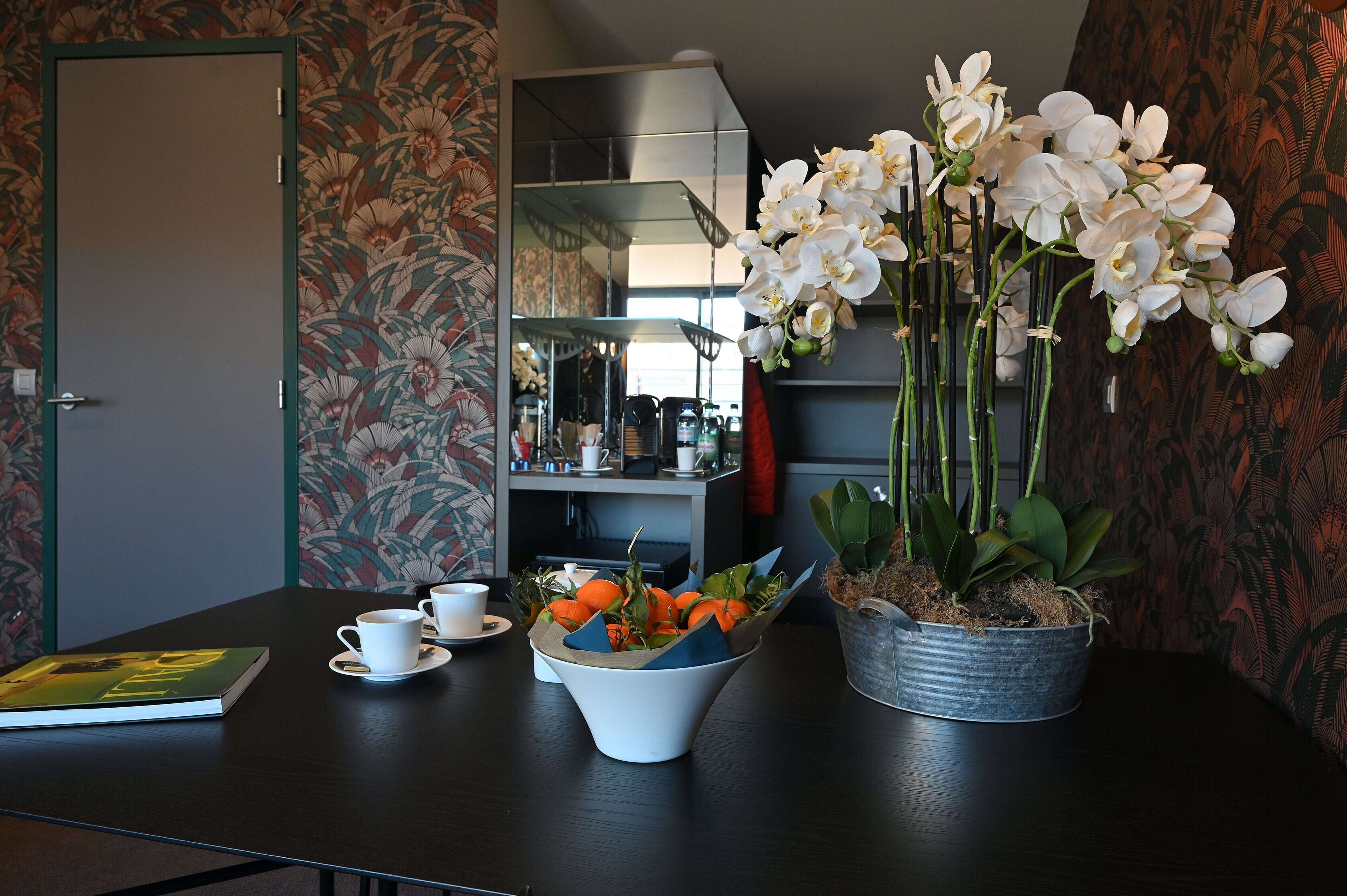 A room with a dark wooden table, a white orchid vase, a potted plant, and a book.