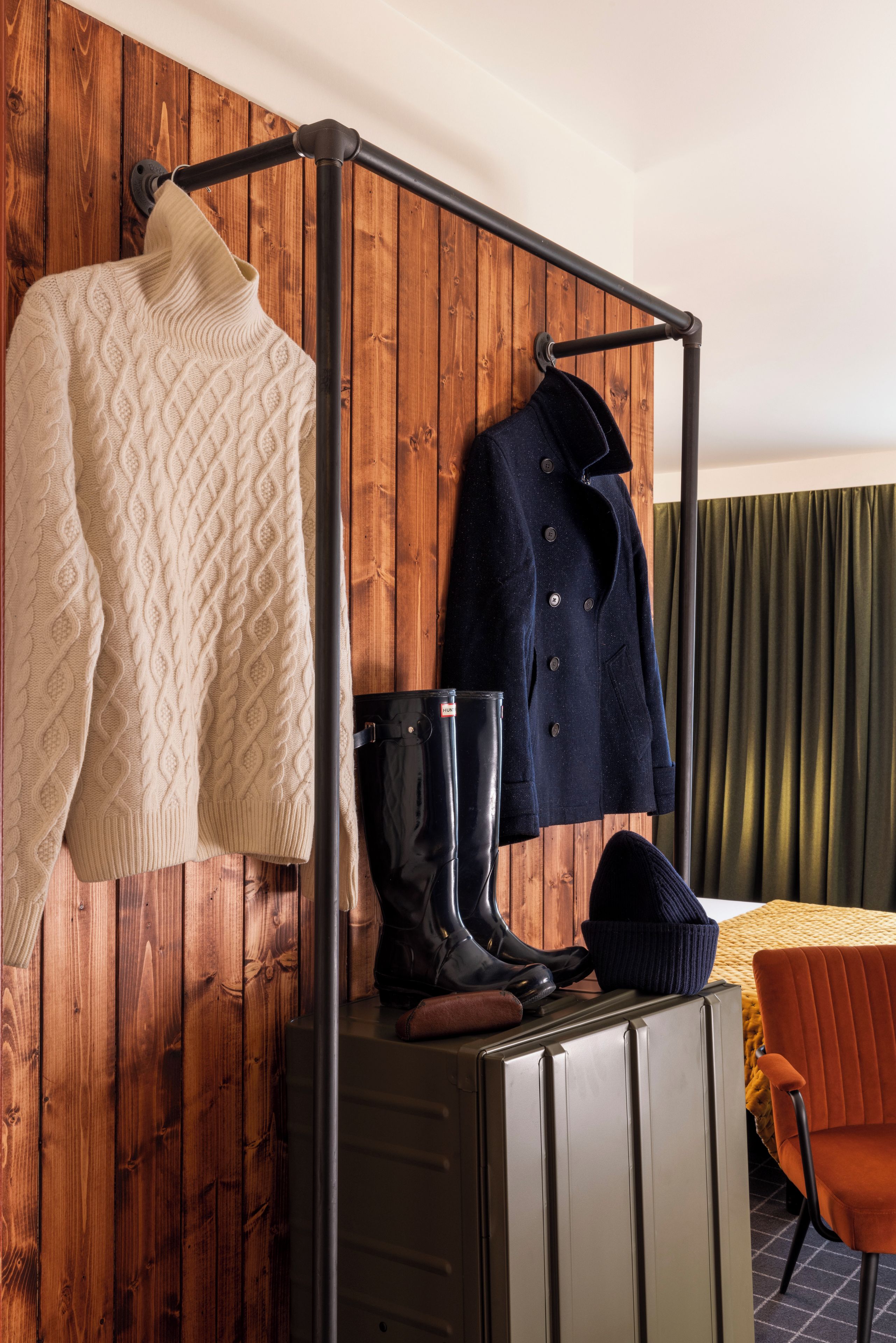 Una habitación con una pared de madera, un perchero metálico con un suéter, botas, un abrigo y una mesa con una silla.