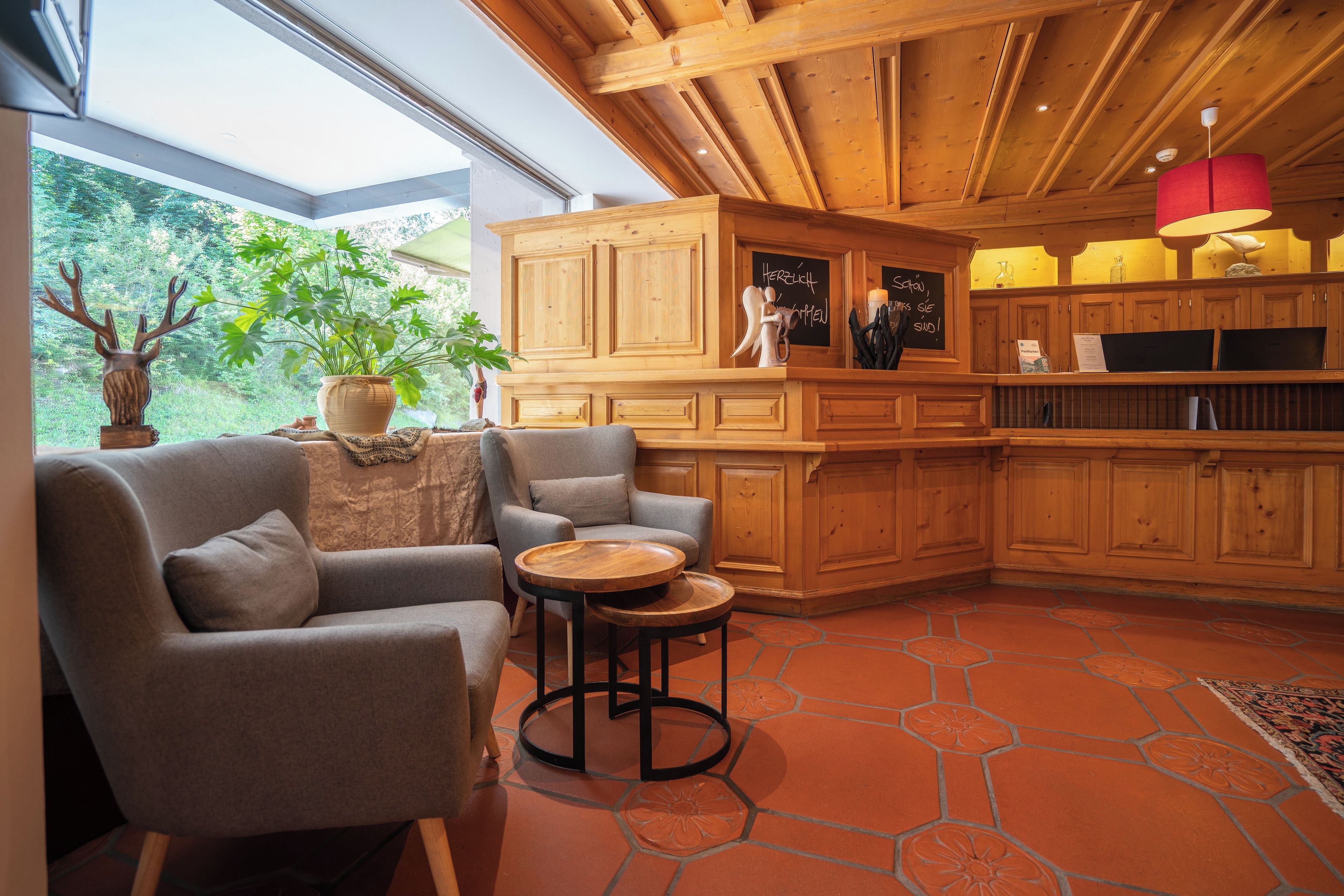 A cozy interior with a wooden ceiling, a seating area with a sofa and armchairs, a small wooden table, and a view of greenery outside.