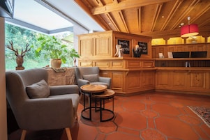 Un interior acogedor con techo de madera, un rincón de estar con sofá y sillones, una pequeña mesa de madera y vistas a la vegetación exterior.